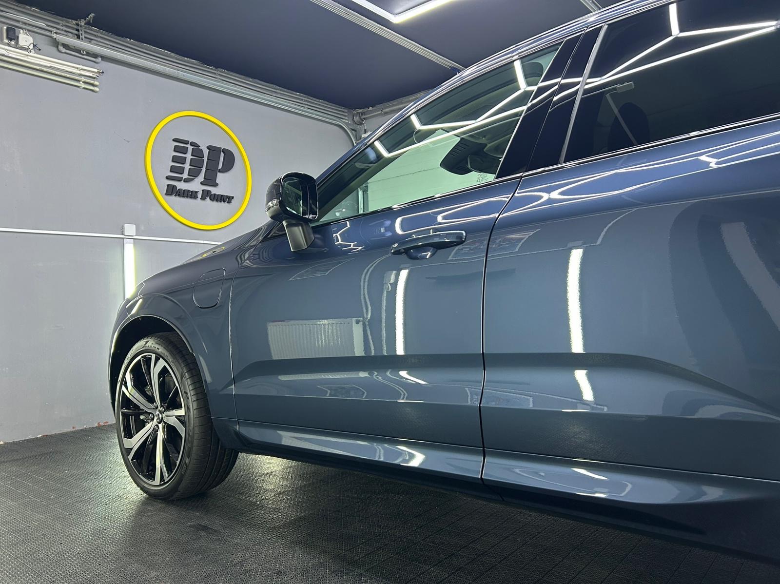 Błyszczący, szary samochód SUV z przyciemnionymi szybami, zaparkowany w dobrze oświetlonym garażu z logo firmy detailerskiej na ścianie.