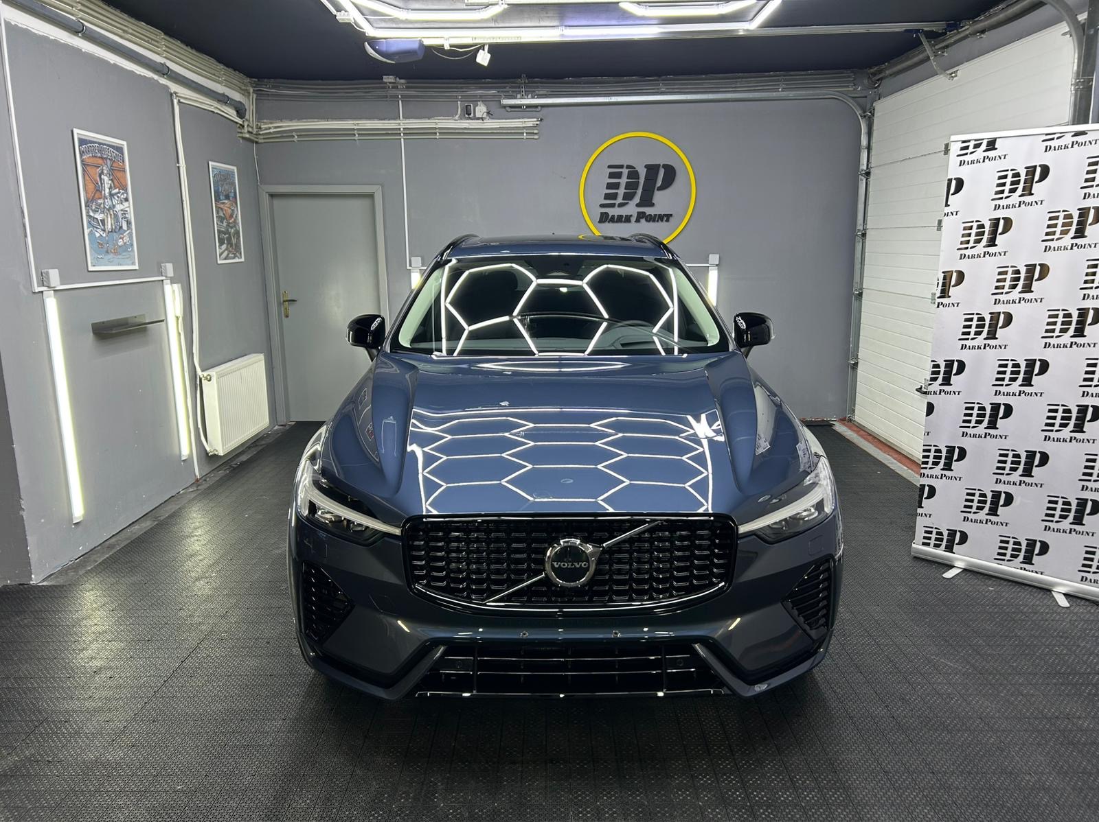 Błękitny SUV Volvo po detailingu, z wyraźnymi odbiciami światła w kształcie plastra miodu na masce, prezentowany na tle szarej ściany z logo firmy detailingowej i plakatem reklamowym.