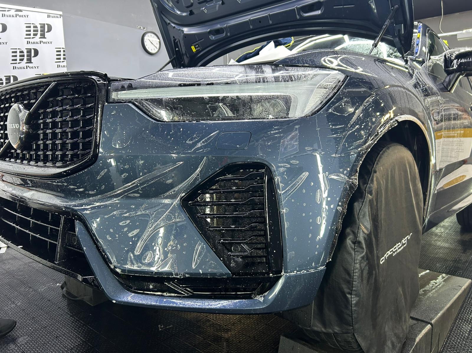 Granatowy samochód Volvo z otwartą maską, częściowo pokryty folią ochronną podczas aplikacji, z widocznym logo Dark Point w tle.