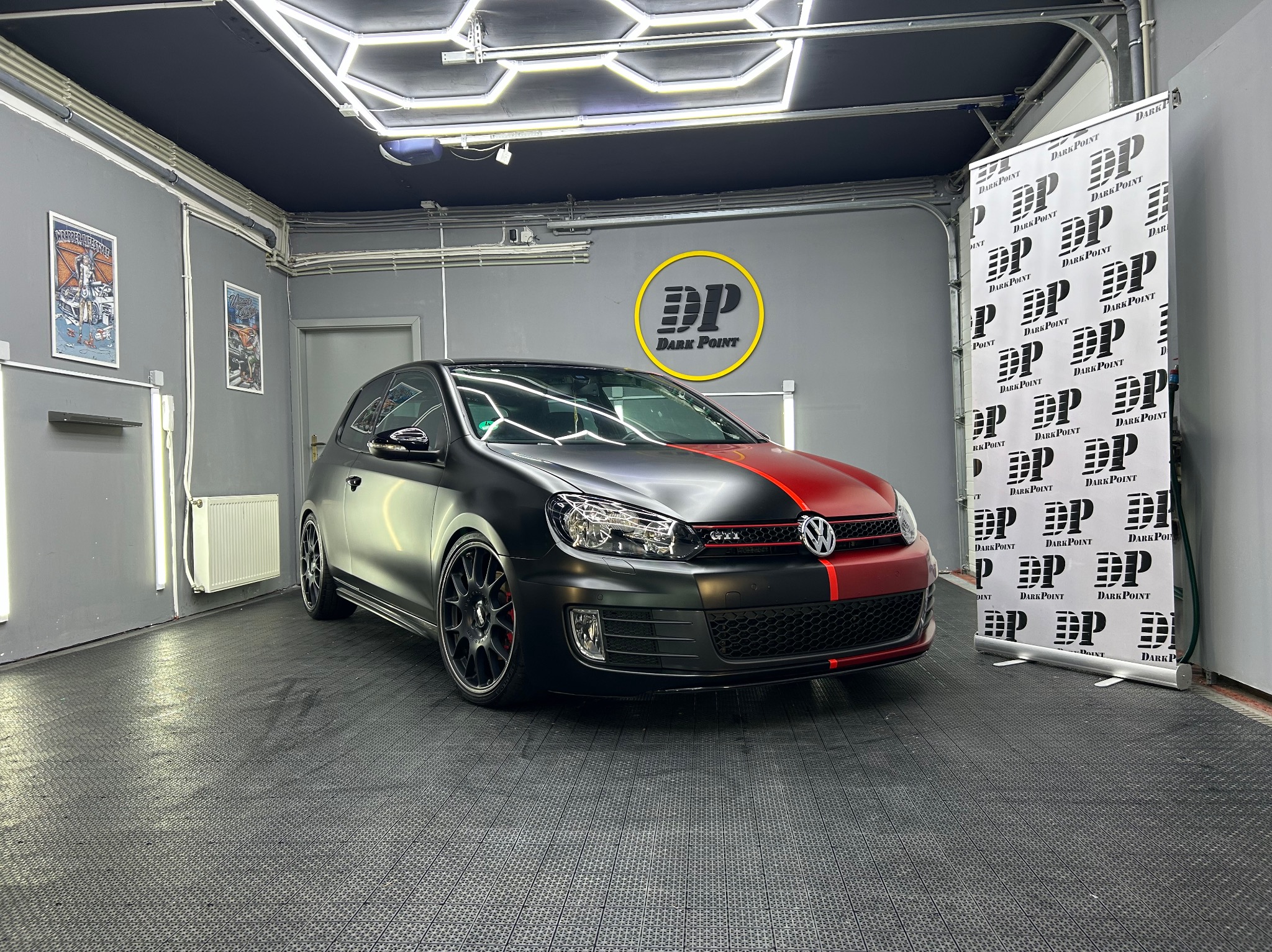 Volkswagen Golf GTI z matową czarną folią i czerwonym pasem, zaparkowany w dobrze oświetlonym studio z logo firmy na ścianie i banerem reklamowym w tle.