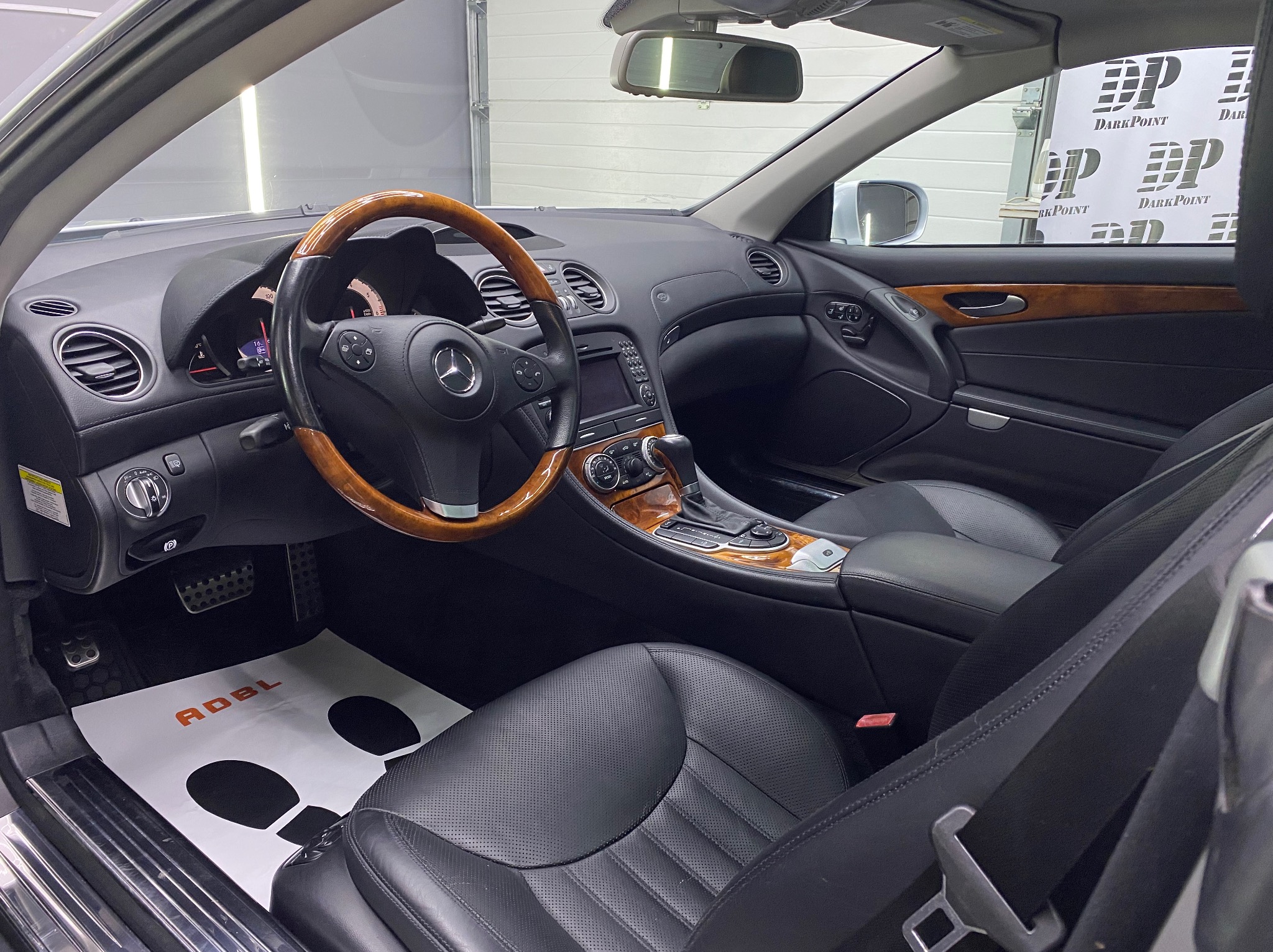 Wnętrze luksusowego samochodu Mercedes SL z drewnianą kierownicą i czarną skórzaną tapicerką po zabiegach pielęgnacyjnych, widoczna mata ochronna na podłodze z logo ADBL oraz logo DarkPoint...