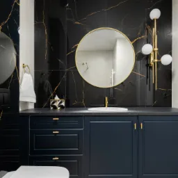 Elegancka łazienka z czarnymi płytkami imitującymi marmur ze złotymi żyłami, okrągłym lustrem w złotej ramie, ciemnoniebieską szafką pod umywalką ze złotymi uchwytami i złotą armaturą, oraz wiszącą...