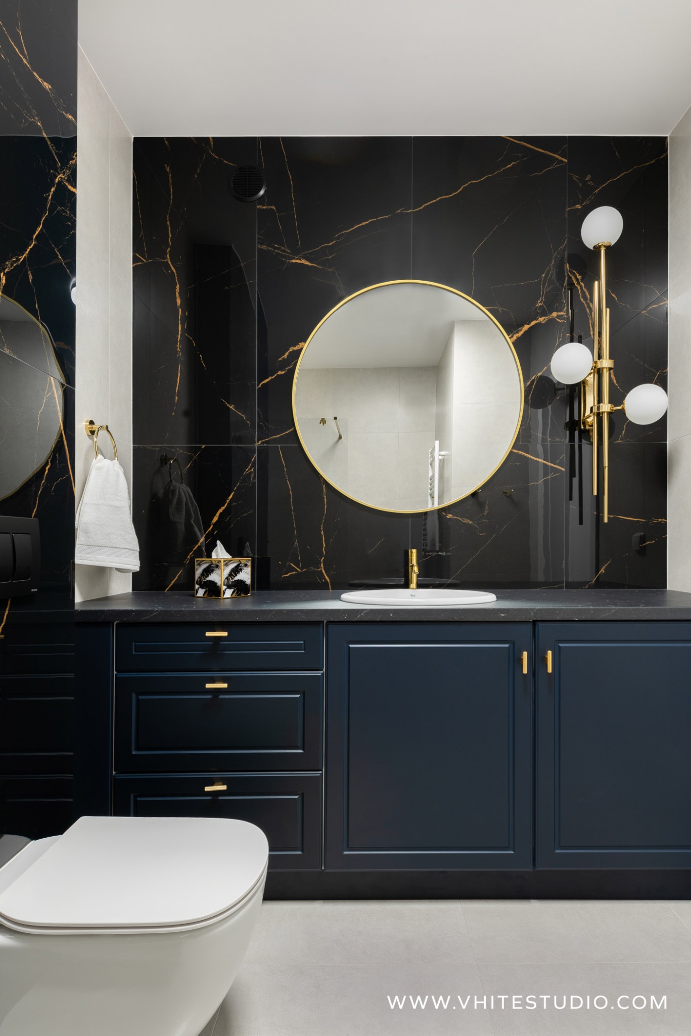Elegancka łazienka z czarnymi płytkami imitującymi marmur ze złotymi żyłami, okrągłym lustrem w złotej ramie, ciemnoniebieską szafką pod umywalką ze złotymi uchwytami i złotą armaturą, oraz wiszącą...