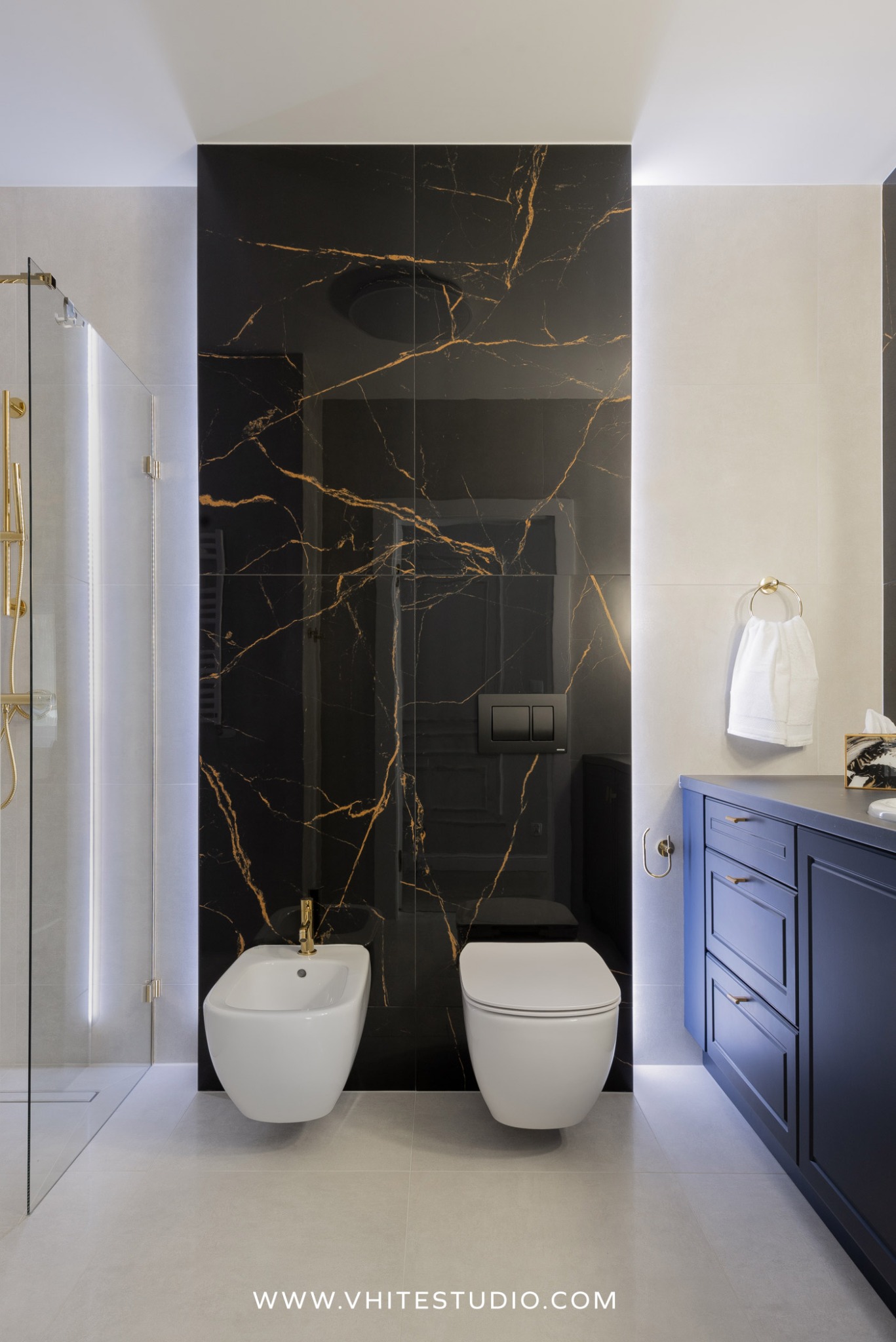 Nowoczesna łazienka z czarną ścianą z efektem marmuru, białym bidetem i toaletą podwieszaną oraz granatową szafką z złotymi uchwytami.