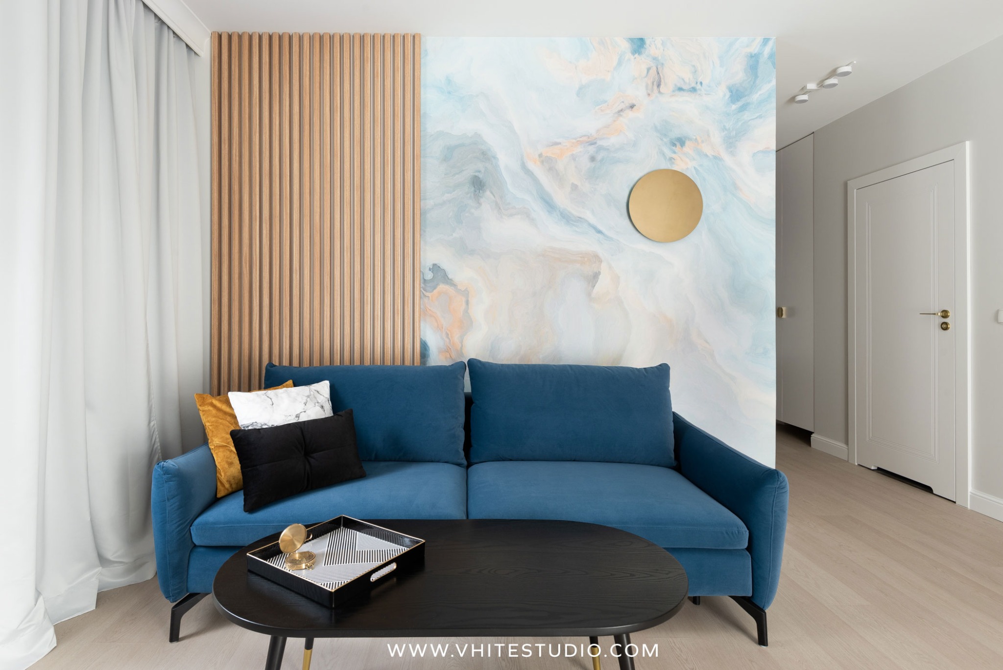 Niebieska sofa z poduszkami na tle ściany z drewnianymi lamelami i abstrakcyjnym wzorem, okrągła złota dekoracja na ścianie, czarny stolik kawowy z geometryczną tacą, jasna podłoga i białe drzwi...
