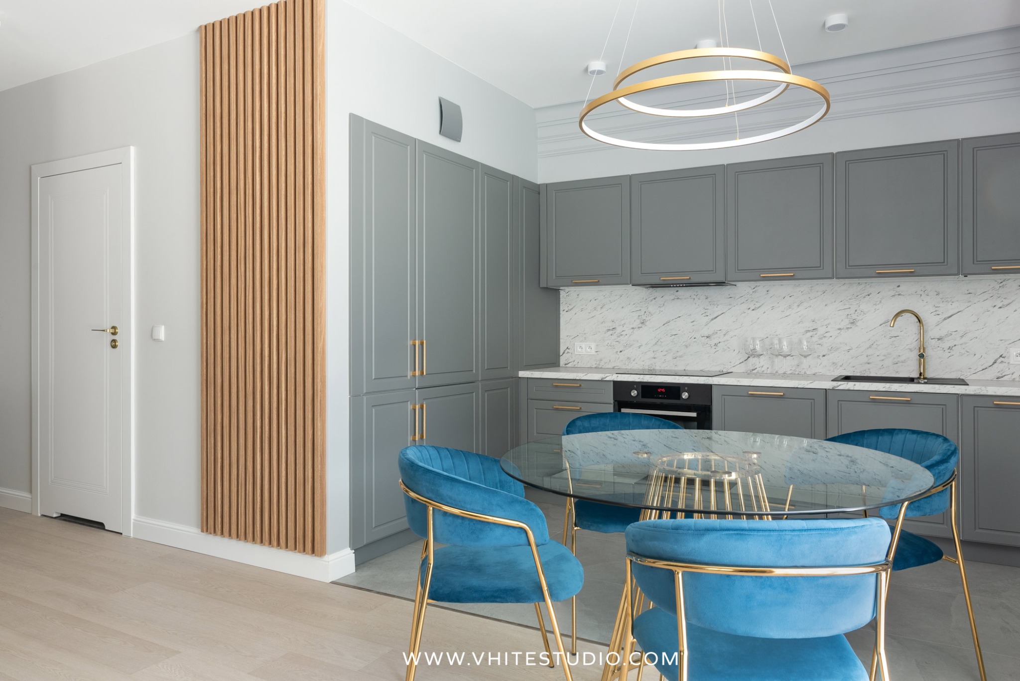 Nowoczesna kuchnia z szarymi szafkami, marmurowym blatem, okrągłym szklanym stołem otoczonym turkusowymi krzesłami z złotymi nogami i dekoracyjnym drewnianym panelem ściennym.