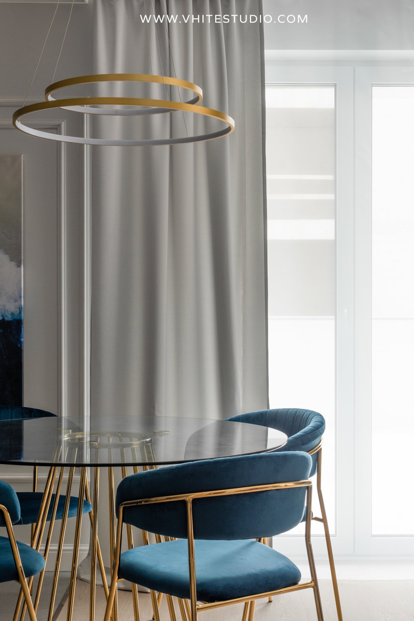 Jadalnia w apartamencie: Szklany stół z metalowymi nogami i aksamitne krzesła w odcieniach granatu, oświetlona dwoma okrągłymi, złotymi lampami wiszącymi.