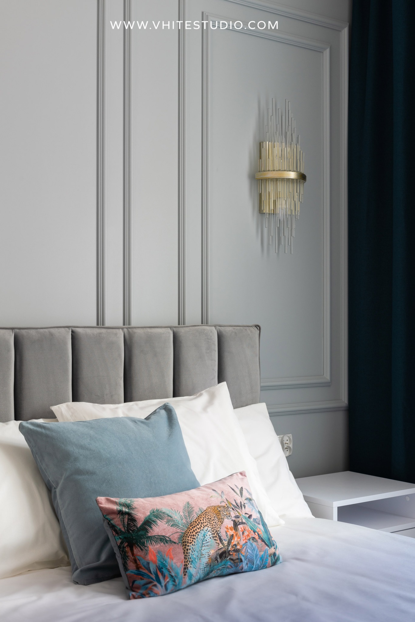 Szczegółowe ujęcie sypialni z szarą tapicerowaną ramą łóżka, dekoracyjnymi poduszkami z motywem tropikalnym i lampą ścienną w stylu art deco na tle szarej ściany z listwami.