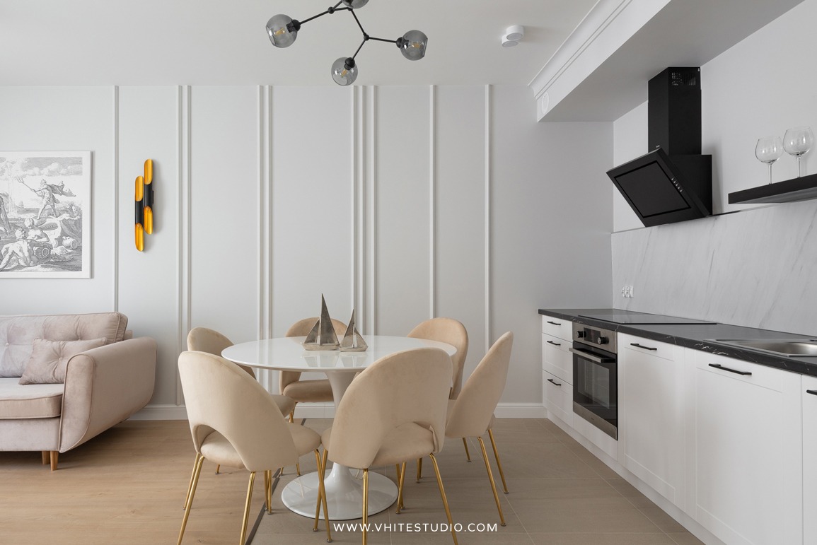 Jasne wnętrze apartamentu z okrągłym stołem, beżowymi krzesłami na złotych nogach, minimalistyczną kuchnią z czarnym blatem i okapem, oraz sofą w tle, oświetlone designerską lampą sufitową i złotym...