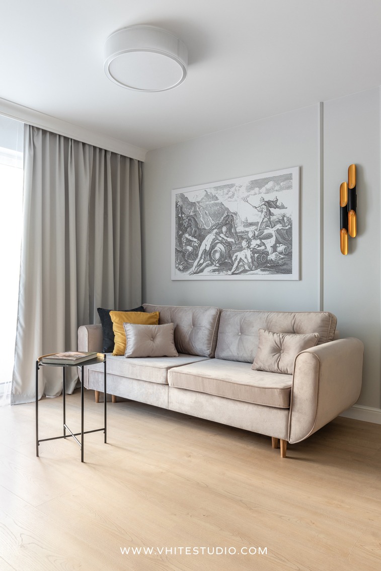 Jasny salon z beżową sofą, czarnym stolikiem pomocniczym i dekoracyjną grafiką na ścianie, oświetlony lampą sufitową i złotym kinkietem.
