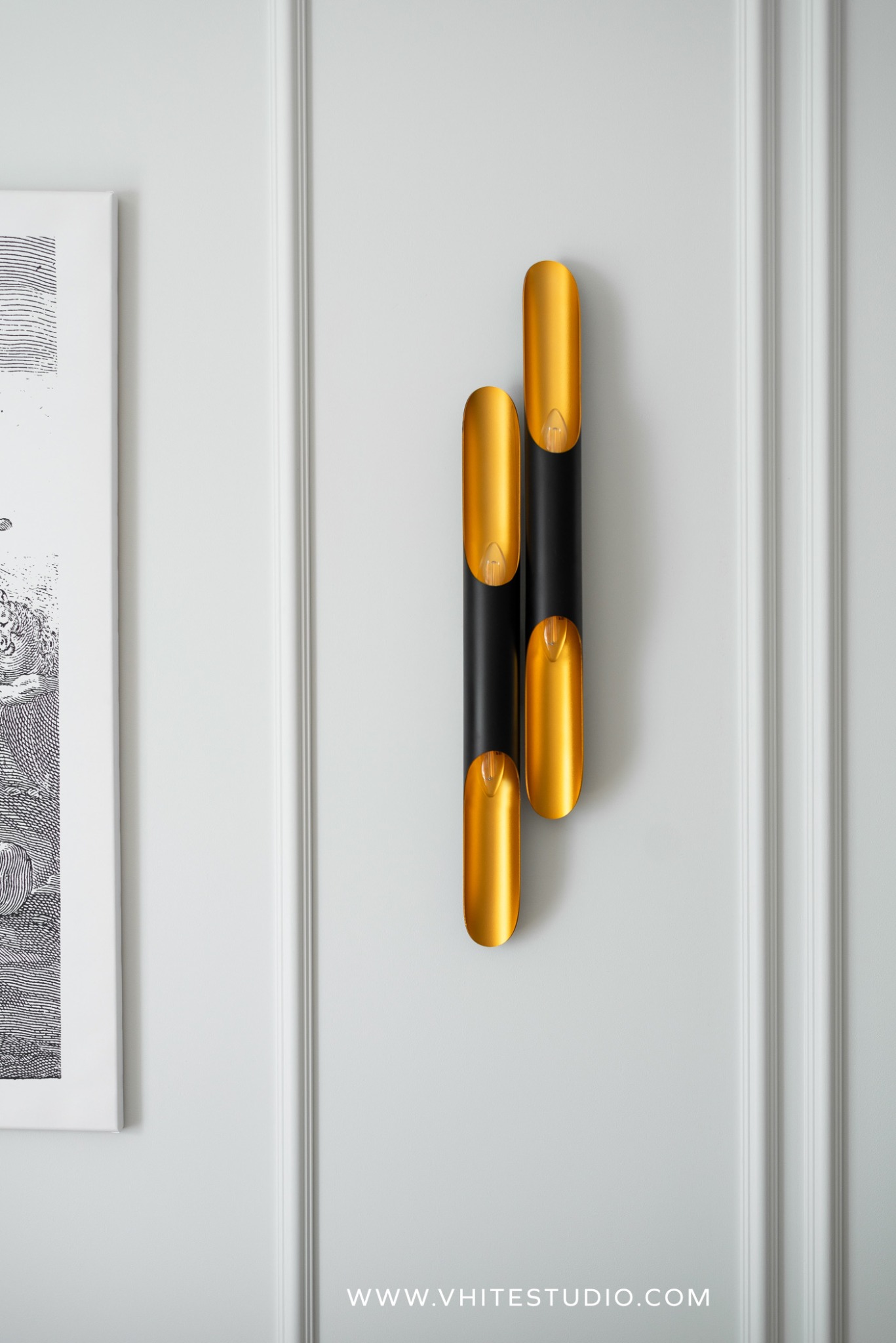 Nowoczesny kinkiet ścienny z elementami w kolorze złota na białej ścianie z dekoracyjną sztukaterią, obok grafika w ramie. Widoczny adres strony internetowej Vhite Studio.