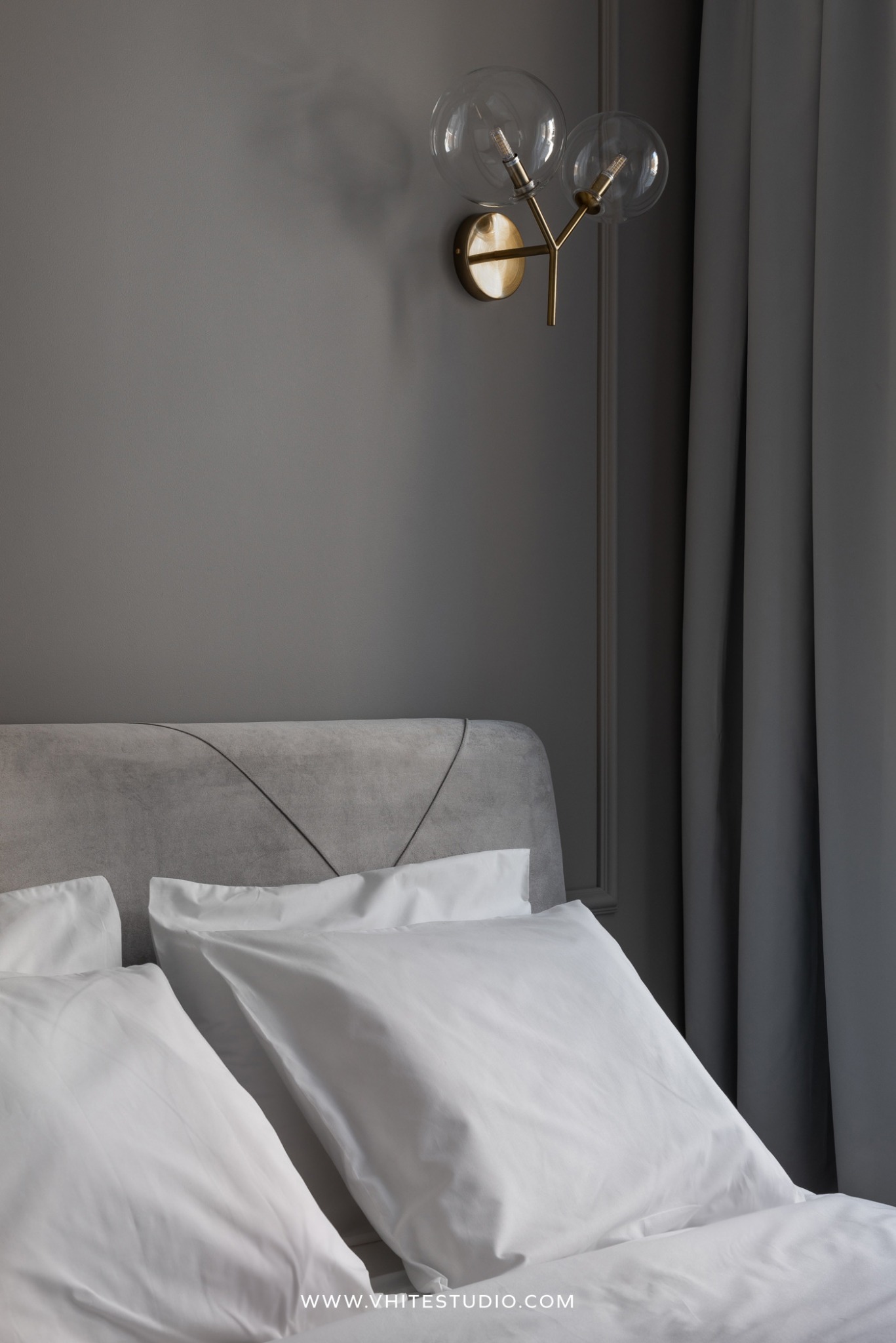 Szczegół sypialni w odcieniach szarości: tapicerowane wezgłowie z geometrycznym przeszyciem, biała pościel, złoty kinkiet z dwoma szklanymi kloszami i dekoracyjna sztukateria ścienna.