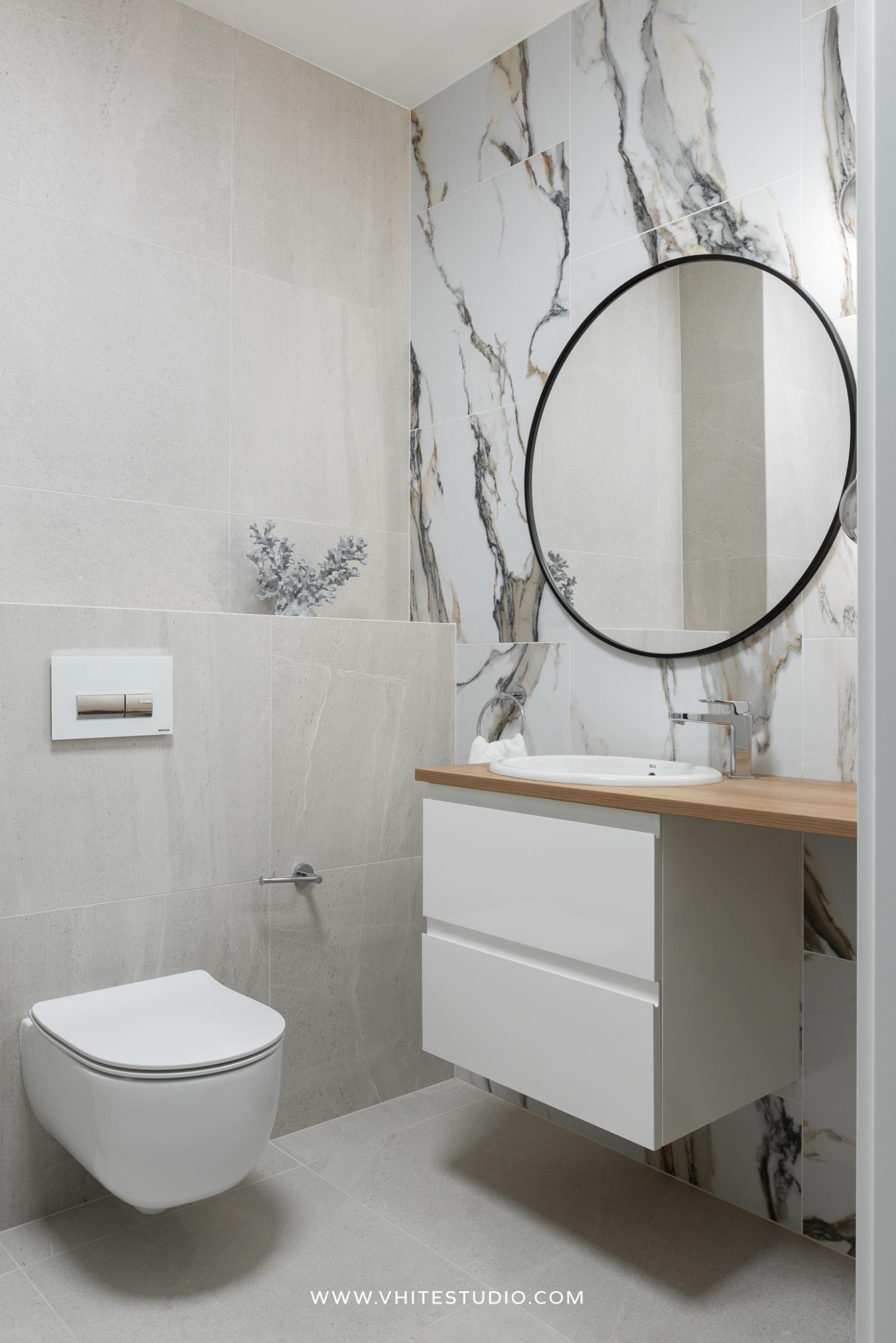Nowoczesna łazienka z wiszącą toaletą, białą szafką pod umywalką z drewnianym blatem, okrągłym lustrem w czarnej ramie i płytkami imitującymi marmur.