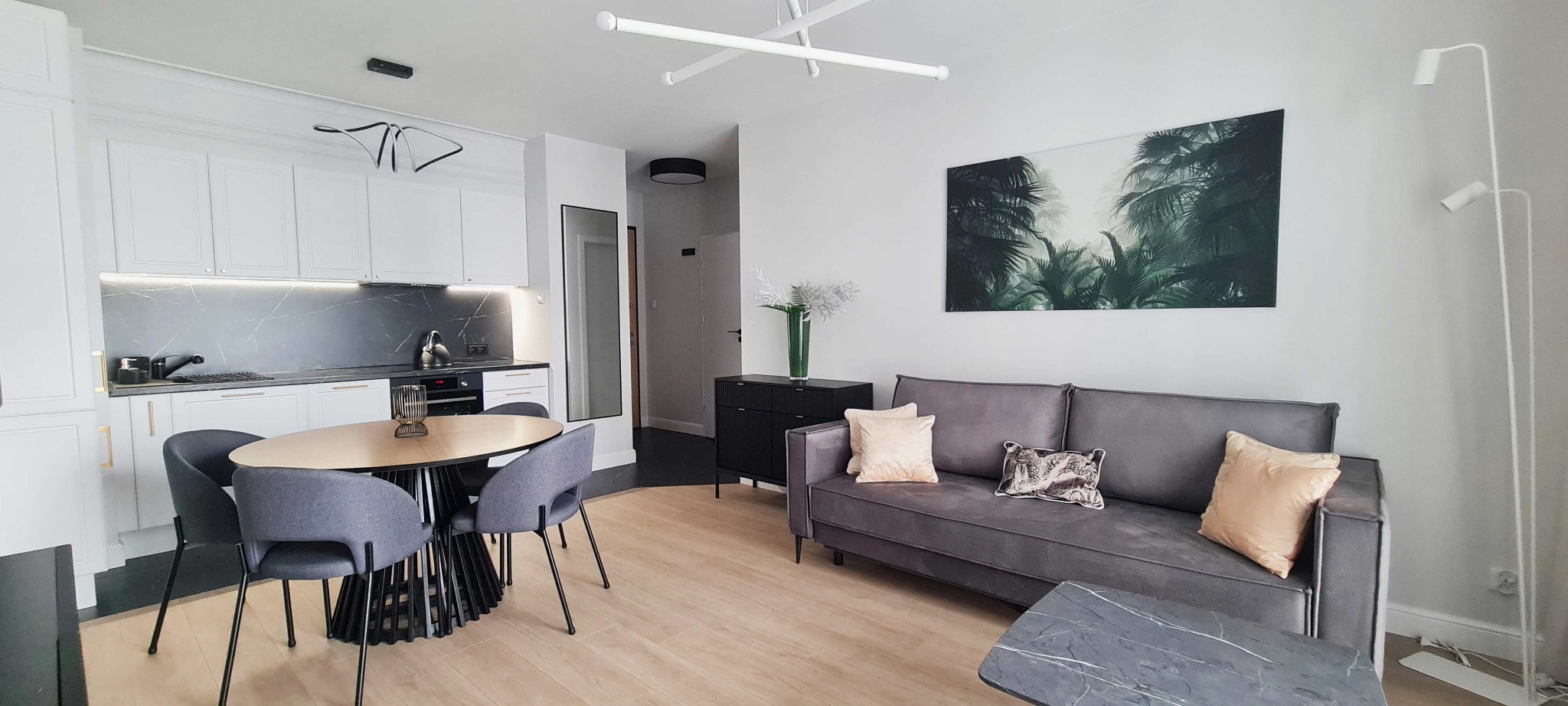 Salon z aneksem kuchennym w nowoczesnym stylu: szara sofa z poduszkami, okrągły stół z krzesłami na tle białej zabudowy kuchennej z czarnym marmurowym panelem.