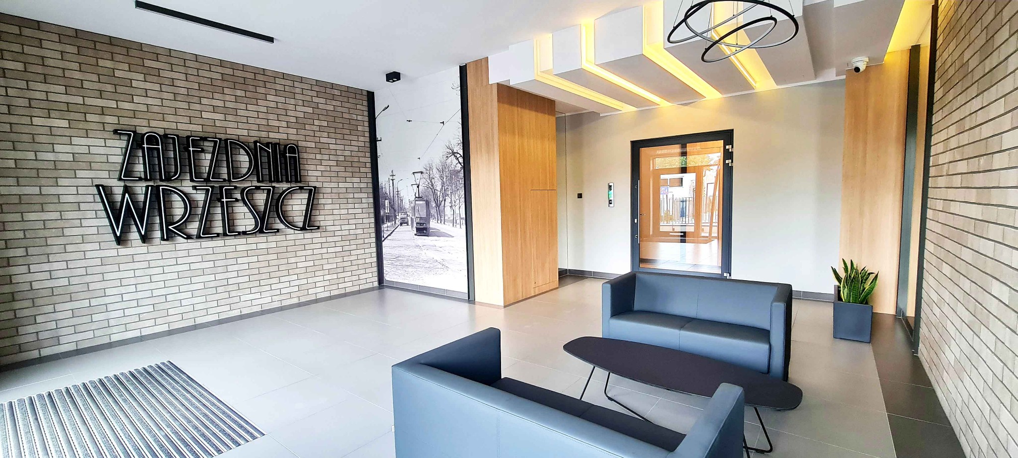 Nowoczesne lobby biurowca z ceglaną ścianą, neonem 'Zajezdnia Wrzeszcz', fototapetą z tramwajem, szarymi sofami i geometrycznym oświetleniem sufitowym.