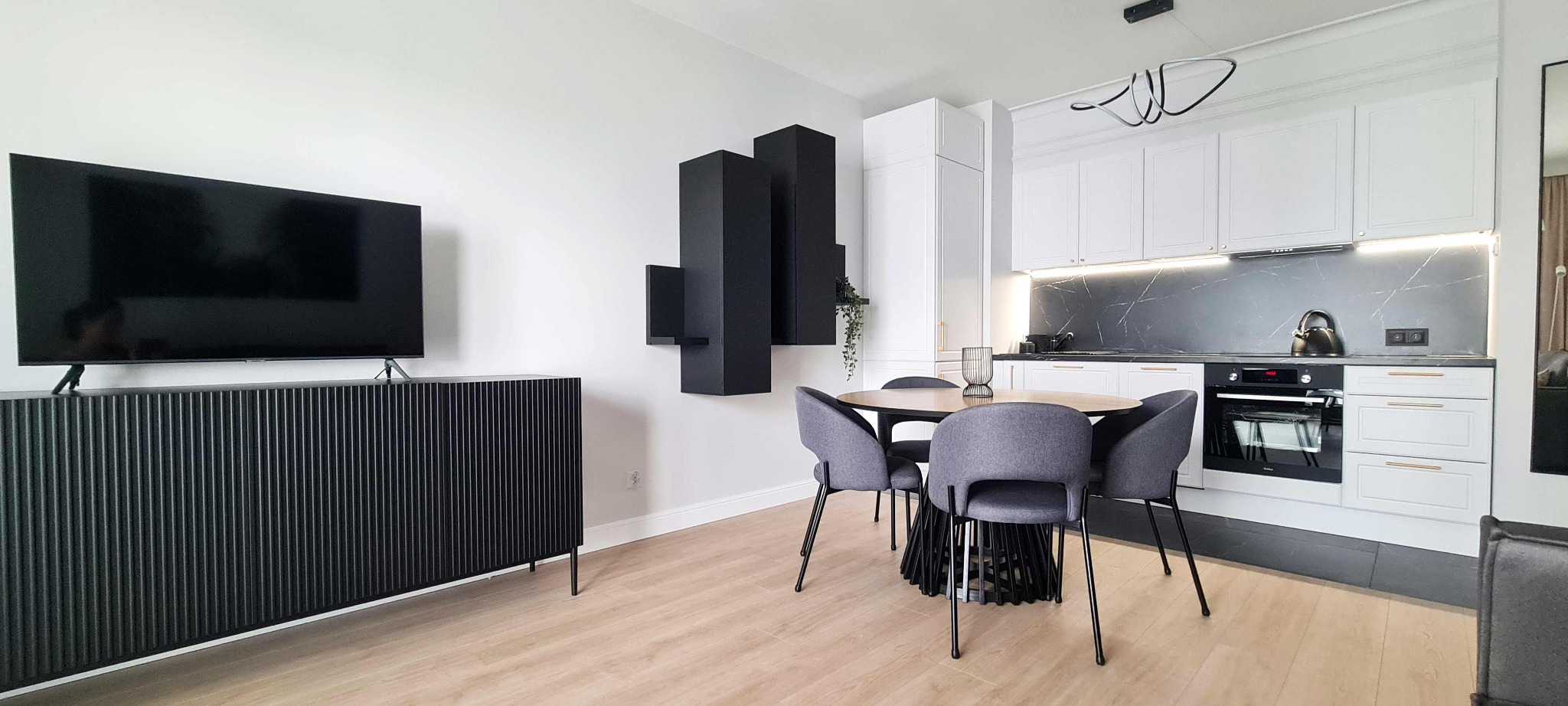 Nowoczesne wnętrze mieszkania z czarnymi akcentami: szafka RTV z lamelami, modułowe półki ścienne, okrągły stół z szarymi krzesłami i biała kuchnia z marmurowym backsplash.