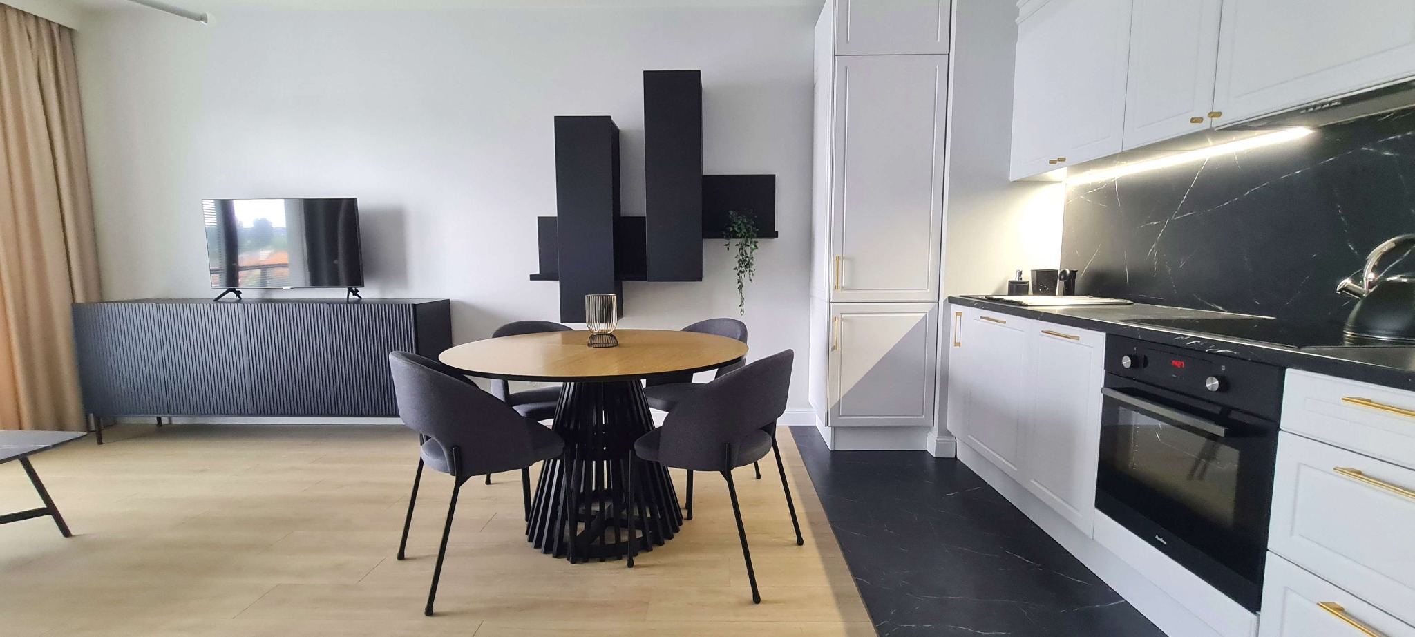 Nowoczesne wnętrze mieszkania z okrągłym stołem z drewnianym blatem, szarymi krzesłami, czarną komodą RTV, wiszącymi czarnymi półkami i białą zabudową kuchenną z czarnym blatem i złotymi uchwytami.