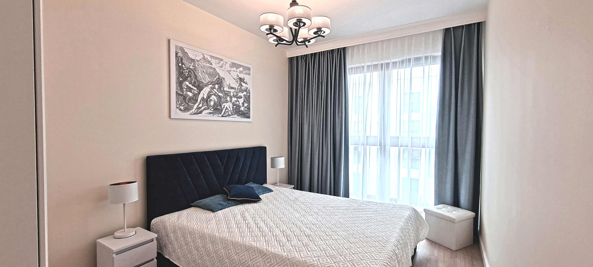 Sypialnia z łóżkiem z ciemnogranatowym zagłówkiem, białą narzutą w geometryczne wzory, szarymi zasłonami i czarno-białym obrazem na ścianie.