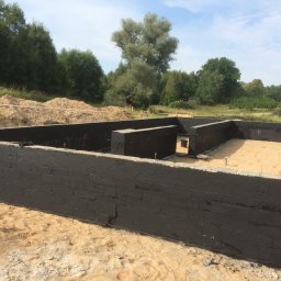 P.H.U. GRONEK ARKADIUSZ GRONKOWSKI - Zabezpieczone fundamenty budynku mieszkalnego, pomalowane na czarno, z widocznymi śladami po pędzlu, na piaszczystym podłożu, w tle drzewa i niebo.