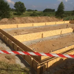 Przygotowanie fundamentów pod budynek: drewniane szalunki zbrojone stalą, widok na wykop z taśmą ostrzegawczą w pierwszym planie, w tle zielone pola i drzewa.