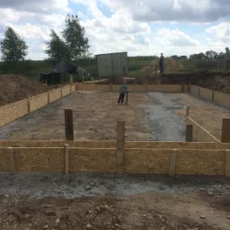 Przygotowanie do wylewki fundamentów: szalunek z płyty OSB, częściowo zalany beton, w tle osoby i sprzęt budowlany przy kopcu ziemi.