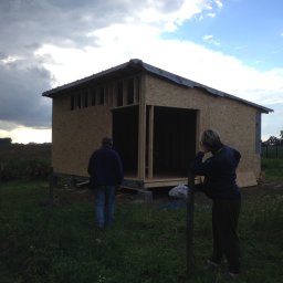 P.H.U. GRONEK ARKADIUSZ GRONKOWSKI - Budowa szkieletowej konstrukcji z płyt OSB na działce, widoczne dwie osoby, jedna wchodzi do środka, druga opiera się o słupek ogrodzenia, w tle pole i zachmurzone niebo.