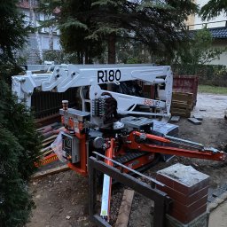 P.H.U. GRONEK ARKADIUSZ GRONKOWSKI - Podnośnik koszowy R180 marki Easylift, ustawiony na ziemi w Morągu, otoczony zielenią drzew i krzewów, z elementami ogrodzenia w pierwszym planie. Na platformie podnośnika widoczny kubek z kawą.