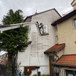 P.H.U. GRONEK ARKADIUSZ GRONKOWSKI - Podnośnik koszowy z pracownikiem czyszczącym elewację domu z białego sidingu, widoczne ślady mycia, obok budynek z elewacją w kolorze piaskowym i czerwoną dachówką, pochmurne niebo.