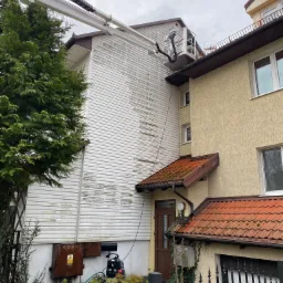 Prace na wysokości: Podnośnik koszowy z pracownikiem podczas czyszczenia sidingu na ścianie budynku, widoczne różnice w czystości powierzchni przed i po myciu, zieleń w tle.