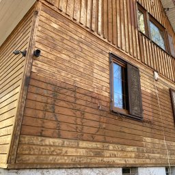 P.H.U. GRONEK ARKADIUSZ GRONKOWSKI - Drewniana elewacja domu z widocznymi śladami zabrudzeń i odchodzącej farby, fragment okna z otwartą okiennicą, kamery monitoringu zamontowane na rogu budynku.