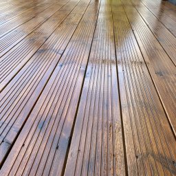 P.H.U. GRONEK ARKADIUSZ GRONKOWSKI - Świeżo zaimpregnowany drewniany taras z widocznymi śladami wilgoci i metalową balustradą w tle, uchwycony pod kątem z bliska.