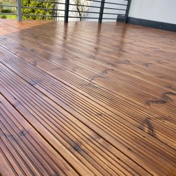 P.H.U. GRONEK ARKADIUSZ GRONKOWSKI - Świeżo polakierowana, brązowa podłoga tarasu z desek w Morągu, z widoczną metalową balustradą i fragmentem elewacji w tle.