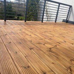 P.H.U. GRONEK ARKADIUSZ GRONKOWSKI - Świeżo zaimpregnowany drewniany taras z metalową barierką na tle zieleni i zabudowań miejskich w oddali, widoczny połysk mokrego drewna.