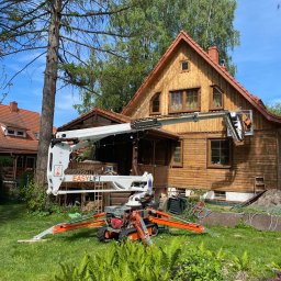P.H.U. GRONEK ARKADIUSZ GRONKOWSKI - Pracownik w pomarańczowym kombinezonie w koszu podnośnika teleskopowego EasyLift, wykonuje prace na drewnianej elewacji domu z brązowym dachem, widok z ogrodu z paprociami i oczkiem wodnym.