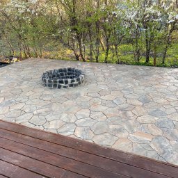 P.H.U. GRONEK ARKADIUSZ GRONKOWSKI - Ułożona z nieregularnych kamieni nawierzchnia tarasu z paleniskiem otoczonym kamieniami, widoczna drewniana podłoga tarasu i kwitnące drzewa w tle.