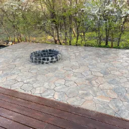 Ułożona z nieregularnych kamieni nawierzchnia tarasu z paleniskiem otoczonym kamieniami, widoczna drewniana podłoga tarasu i kwitnące drzewa w tle.