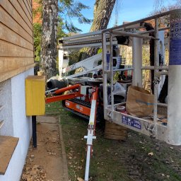 P.H.U. GRONEK ARKADIUSZ GRONKOWSKI - Pomarańczowy podnośnik koszowy EasyLift ustawiony obok drewnianej elewacji budynku, widoczna żółta skrzynka na elewacji i torba Castorama w koszu podnośnika.