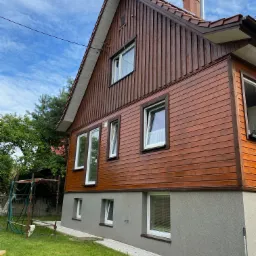 Odnowiona drewniana elewacja domu jednorodzinnego z widocznymi nowymi oknami PCV, szary cokół, trawnik i rusztowanie w tle.