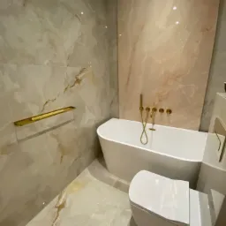 Elegancka łazienka z marmurowymi płytkami w odcieniach beżu i złota, wolnostojąca wanna z baterią podtynkową, wisząca toaleta z deską wolnoopadającą i pozłacany wieszak na ręczniki.