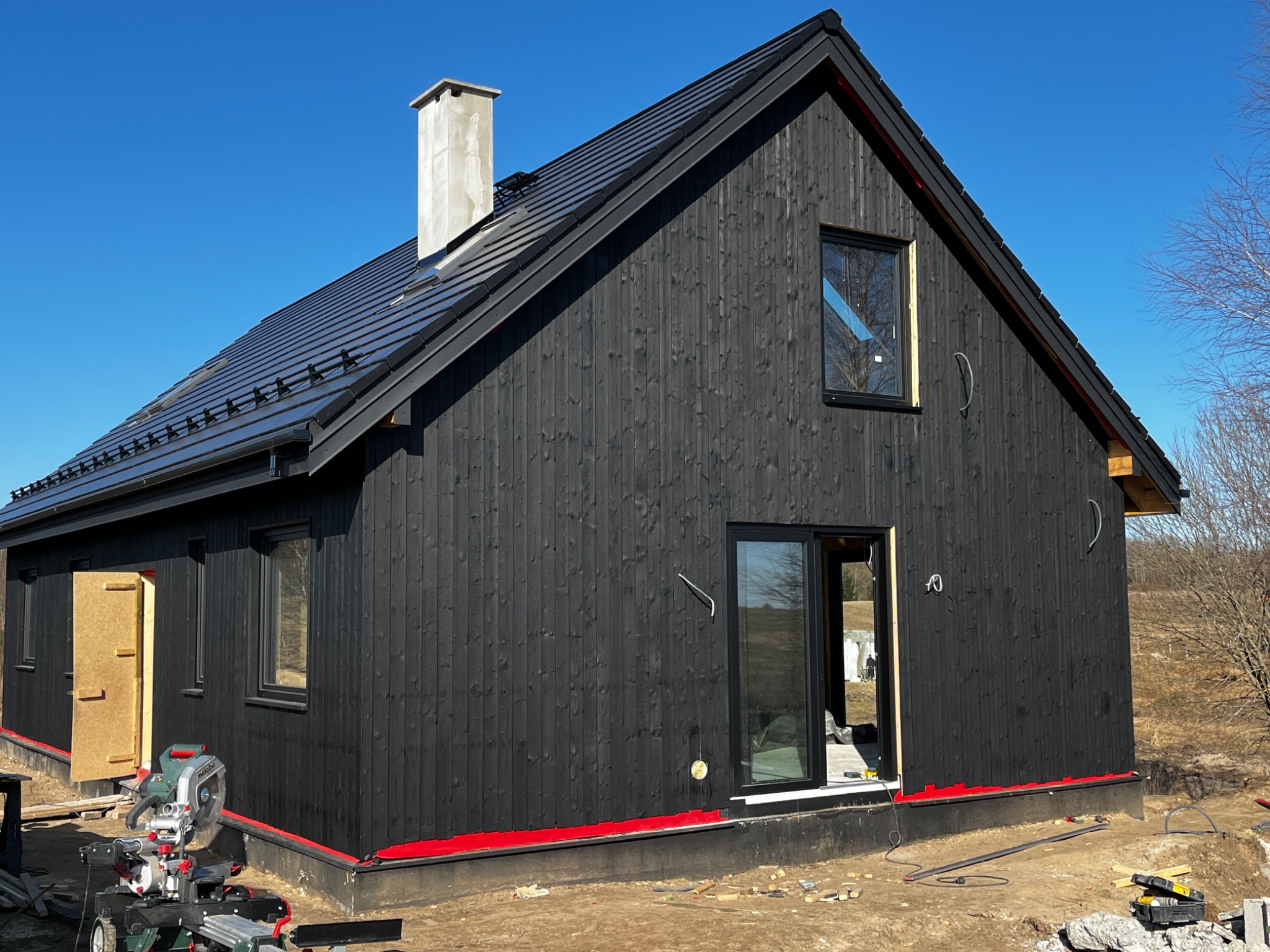 Czarny, drewniany dom z ciemną dachówką w trakcie budowy, z widoczną piłą do drewna i otwartymi drzwiami. Czerwona izolacja przy fundamencie. Błękitne niebo w tle.