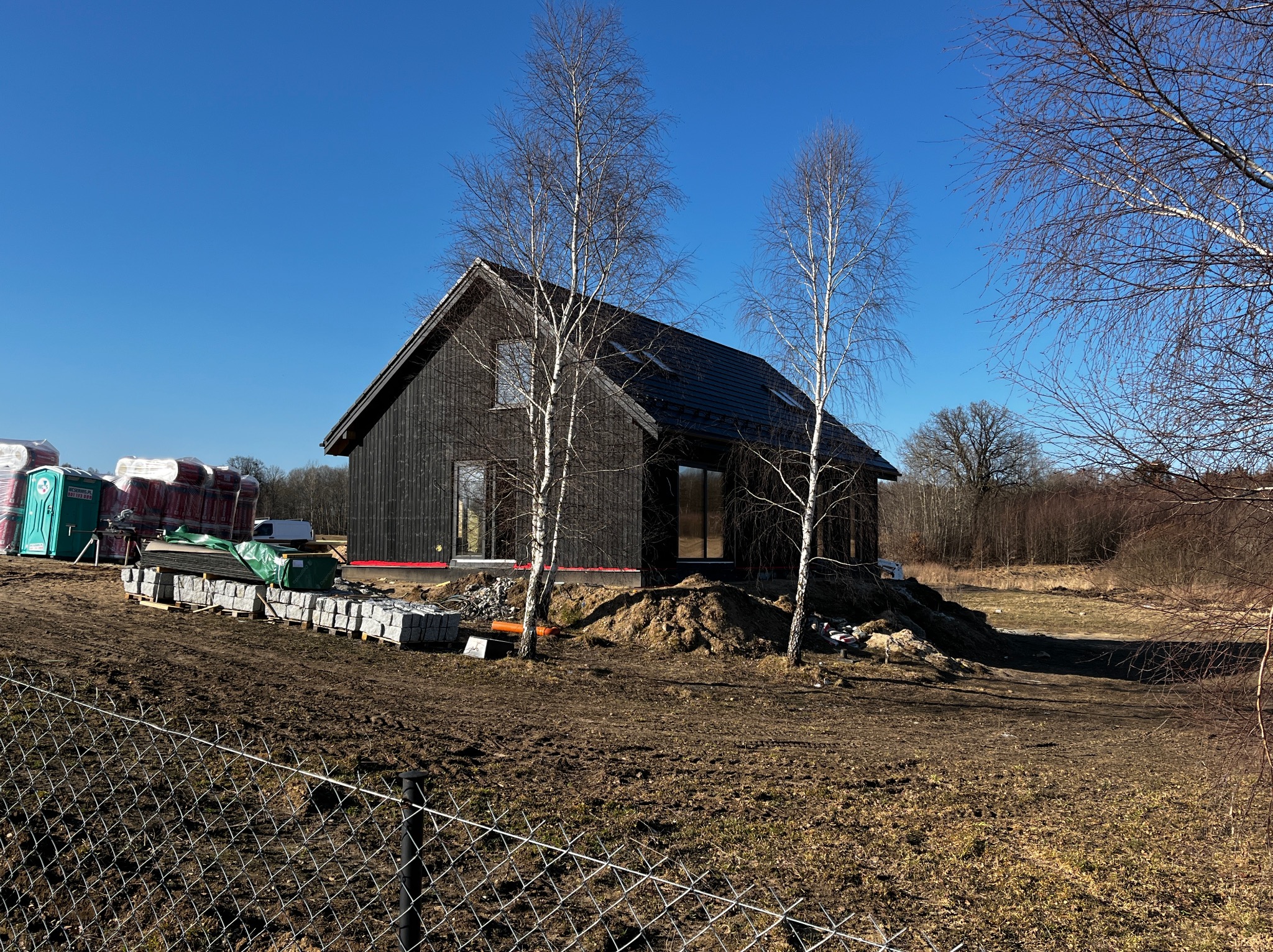 Nowoczesny, ciemny dom z dwuspadowym dachem w trakcie budowy, z widocznymi materiałami budowlanymi i tymczasową toaletą na działce. Błękitne niebo w tle.