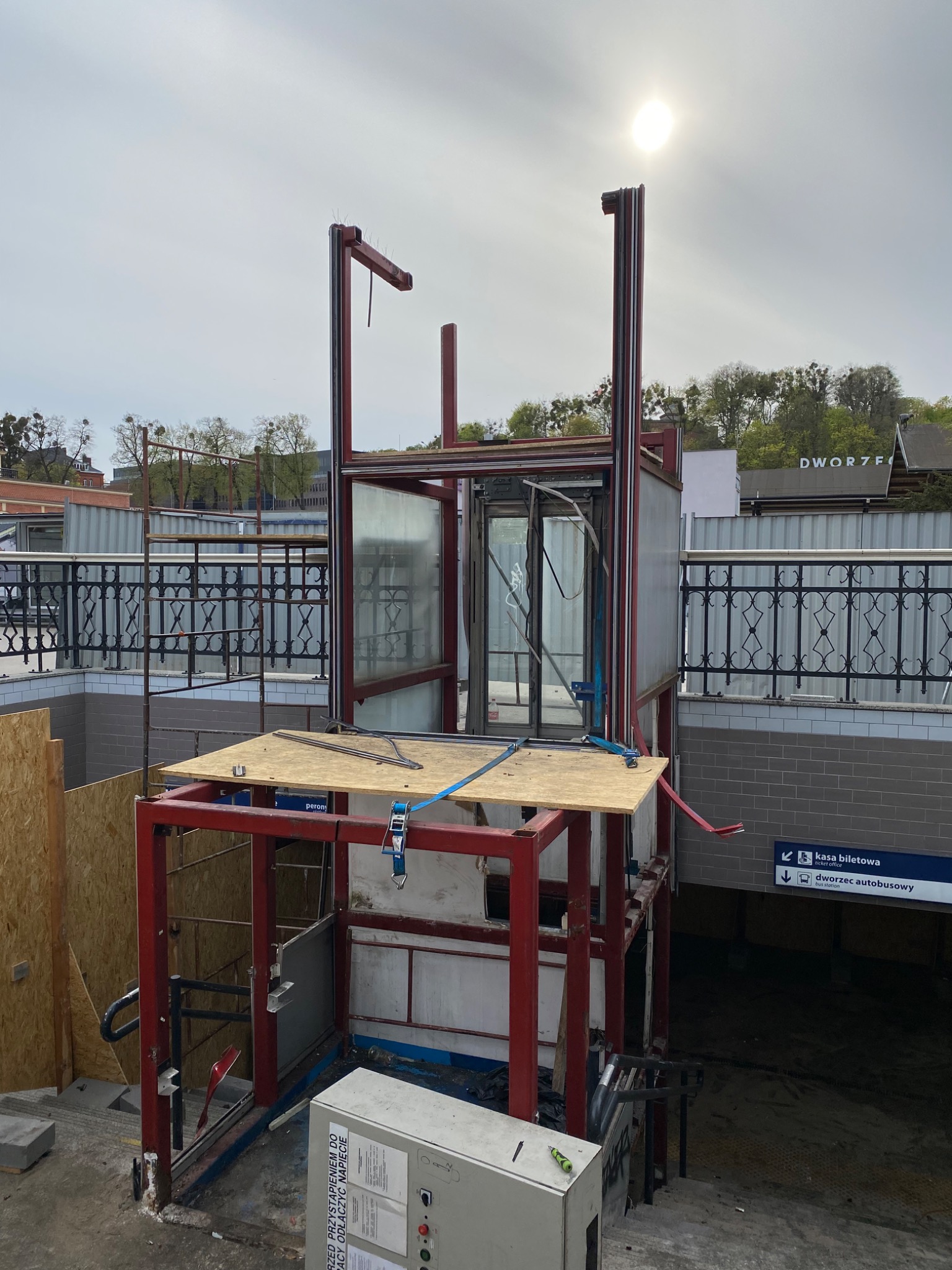 Montaż platformy windowej dla osób niepełnosprawnych przy wejściu do dworca, widoczne stalowe ramy konstrukcji, częściowo przeszklona kabina, płyta OSB zabezpieczona pasami, w tle balustrada...