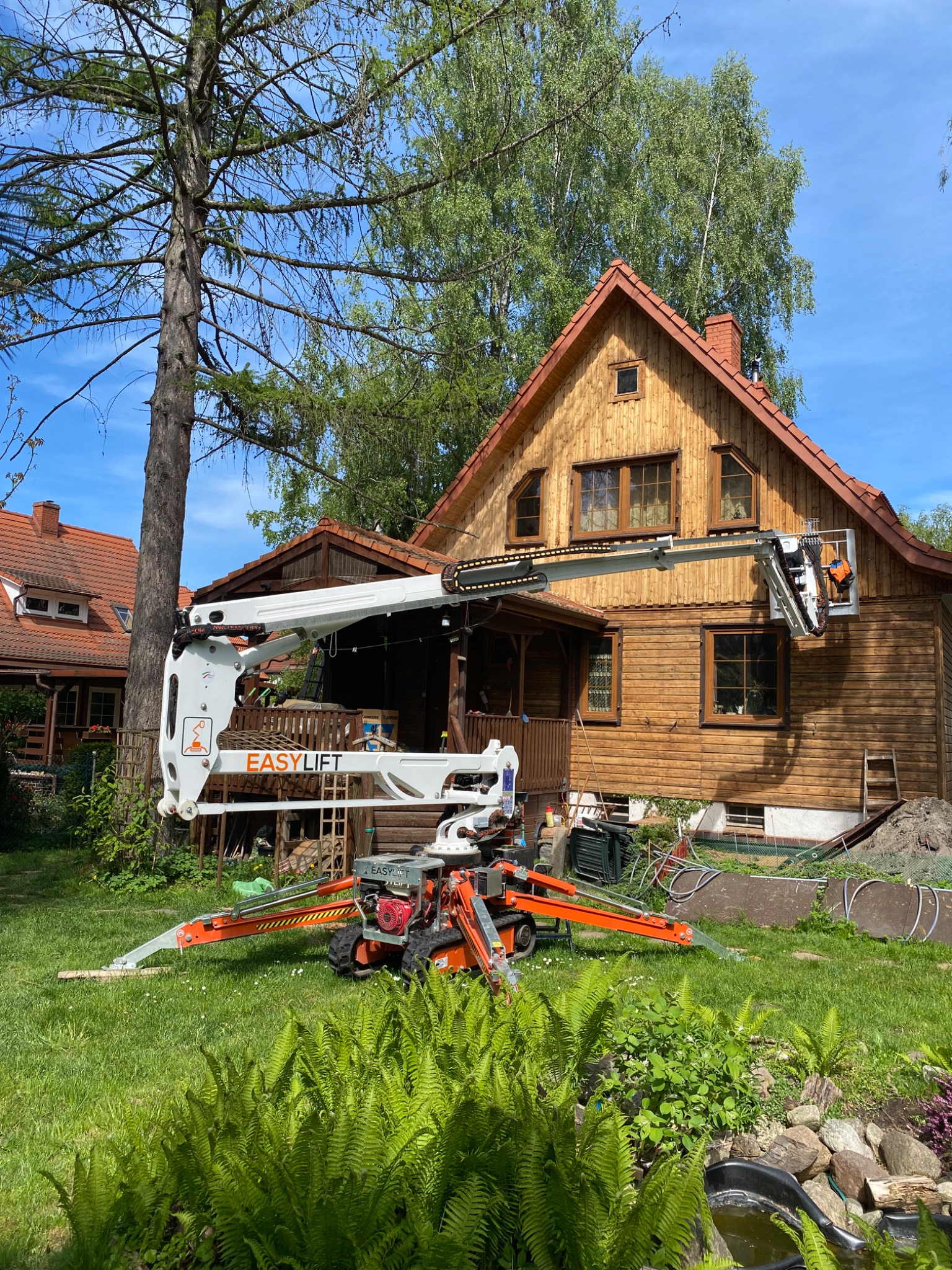 Pracownik w pomarańczowym kombinezonie w koszu podnośnika teleskopowego EasyLift, wykonuje prace na drewnianej elewacji domu z brązowym dachem, widok z ogrodu z paprociami i oczkiem wodnym.