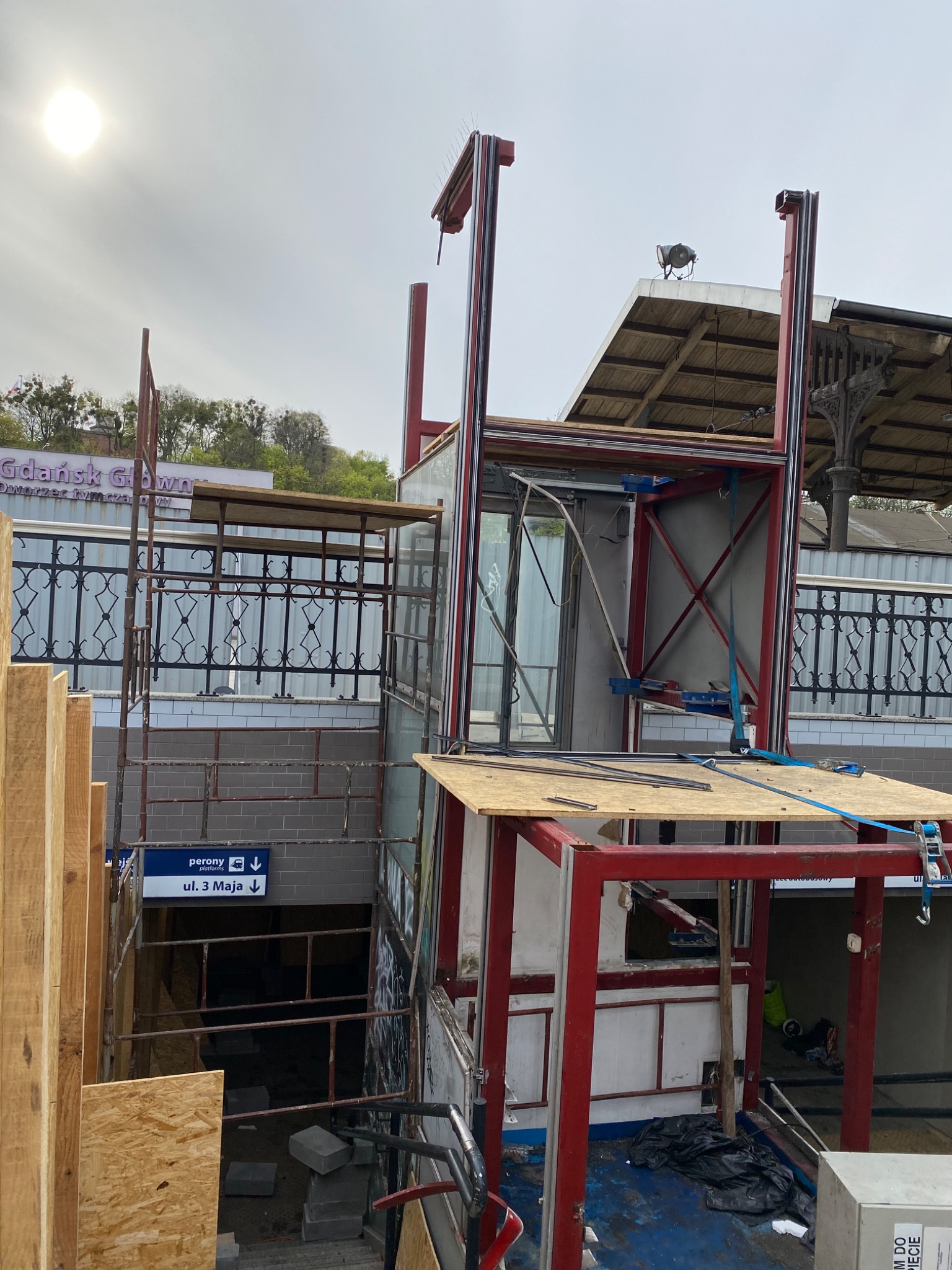 Montaż konstrukcji windy zewnętrznej przy wejściu do stacji kolejowej Gdańsk Główny, widoczne rusztowania, elementy stalowe, drewniane płyty, oznakowanie peronów i zejście na ul. 3 Maja.