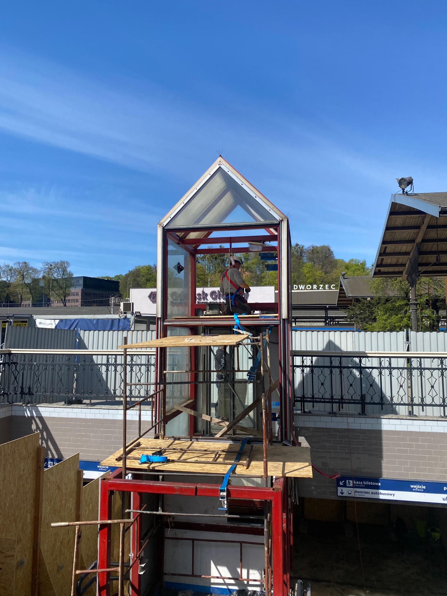 Montaż przeszklonej windy zewnętrznej przy wejściu do dworca autobusowego, pracownik w uprzęży na rusztowaniu, błękitne niebo.