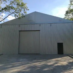 Gotowa hala magazynowa z szarej blachy trapezowej, z dużymi przesuwnymi drzwiami i dodatkowym wejściem, widoczna tabliczka informacyjna na górze ściany szczytowej.