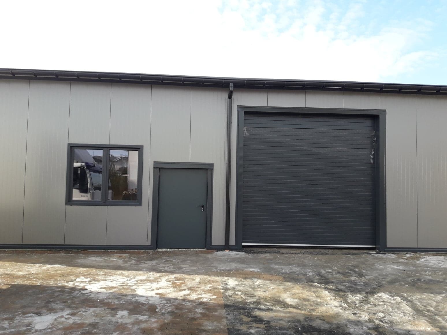 Zewnętrzny widok hali przemysłowej z szarej płyty warstwowej z bramą segmentową i oknem, na ziemi resztki śniegu.