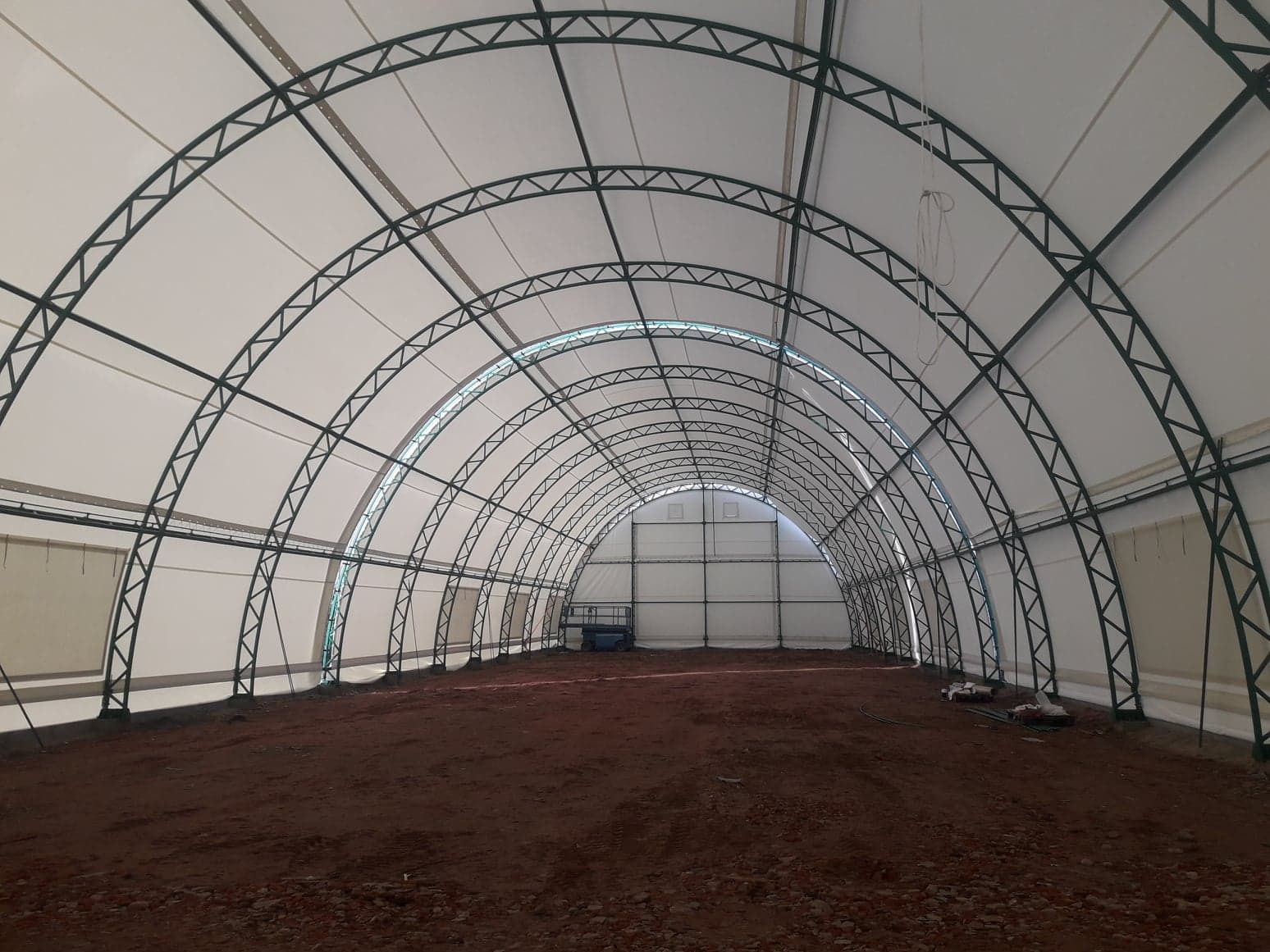 Wnętrze hali z konstrukcją stalową łukową i poszyciem z białej membrany, ziemia na podłożu, niebieski podnośnik w tle.