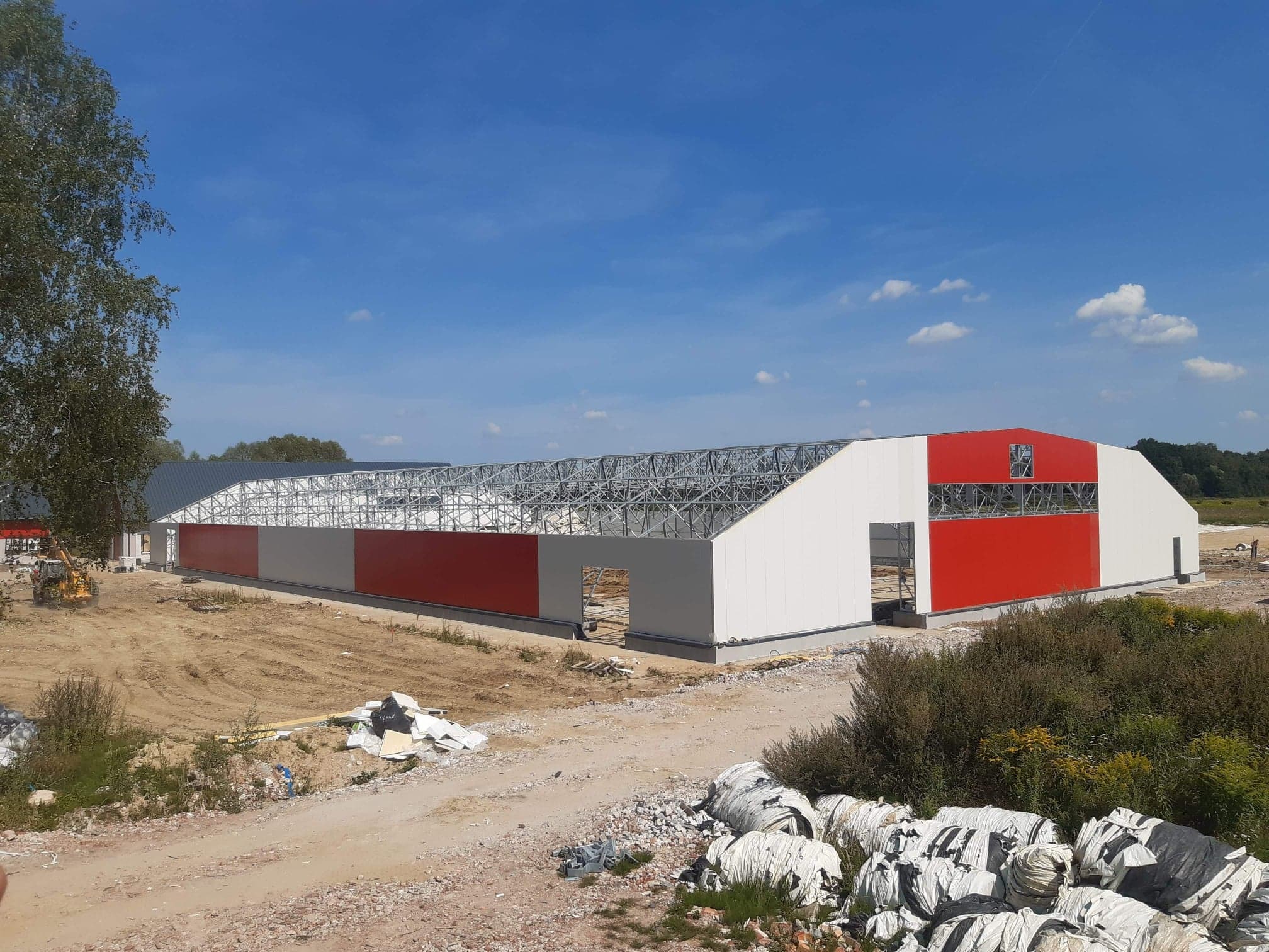 Wznoszona hala stalowa z częściowo zamontowanymi płytami warstwowymi, widoczna konstrukcja dachu, czerwono-białe panele elewacyjne, teren budowy z gruzem i roślinnością.