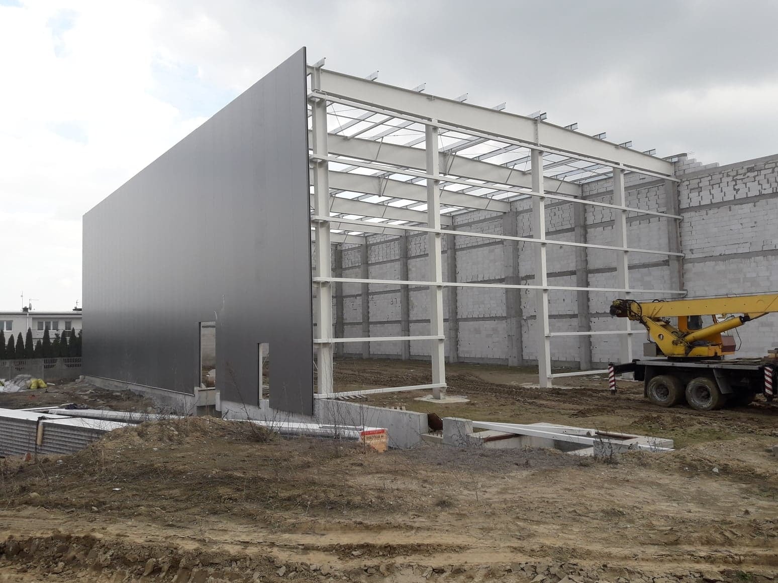 Konstrukcja stalowa hali w trakcie budowy, częściowo obłożona szarymi płytami warstwowymi, na placu budowy stoi żółty dźwig.